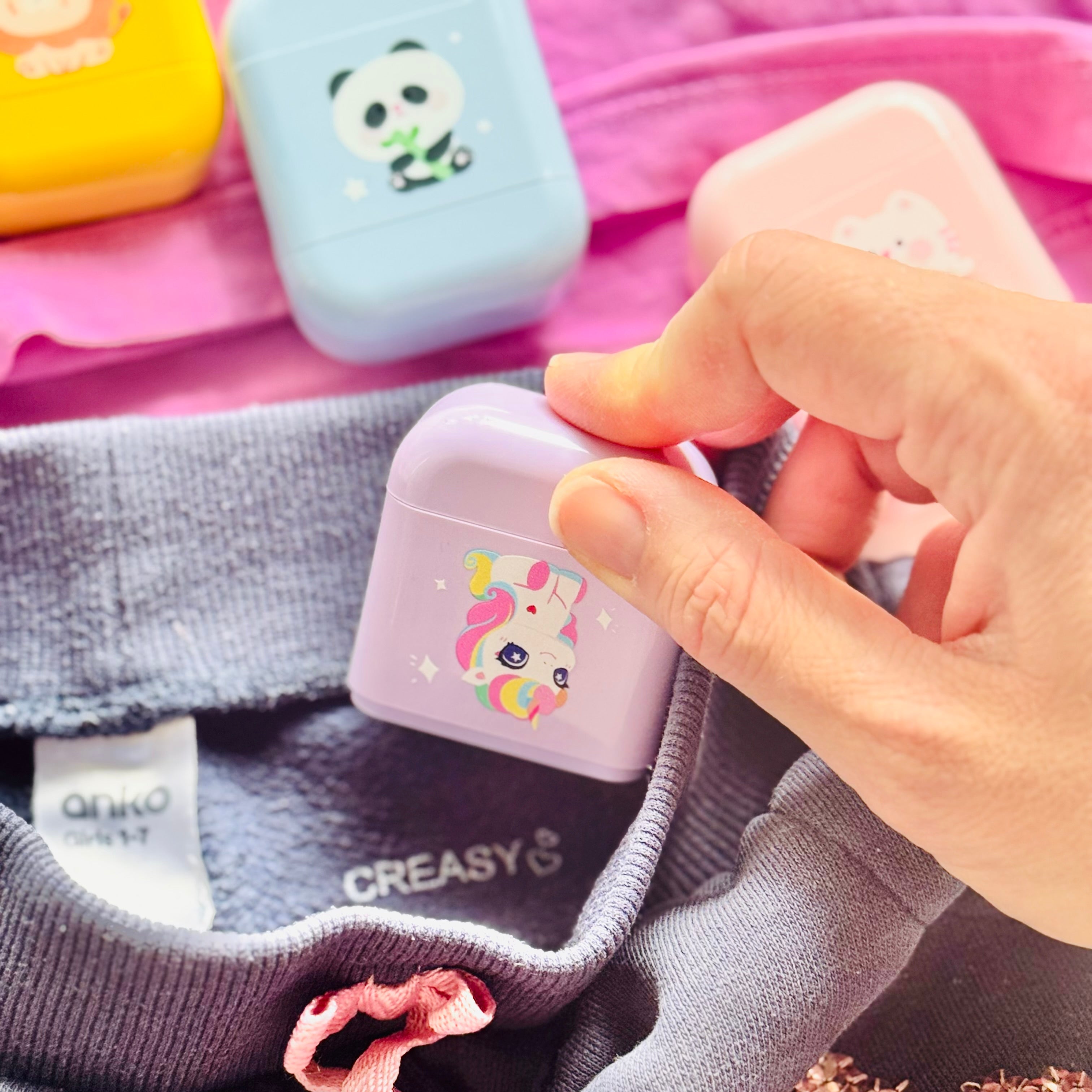 A hand holds a purple name stamp over a childs jumper, a blue and a yellow name stamp in the background.