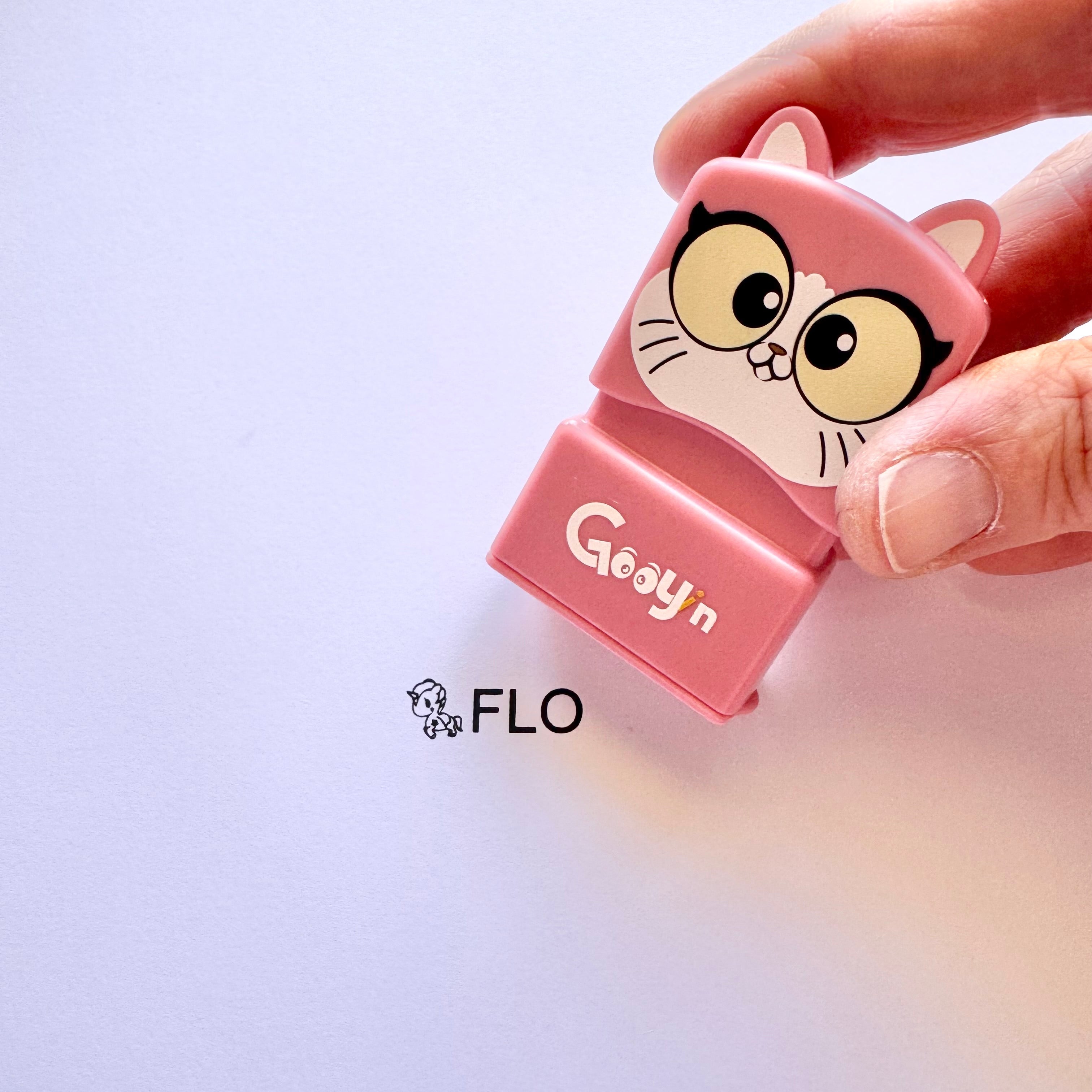 A person holding a cat name stamp with 'Gooyin' printed on the front above a sheet of paper with 'FLO' printed on it with a unicorn at the start