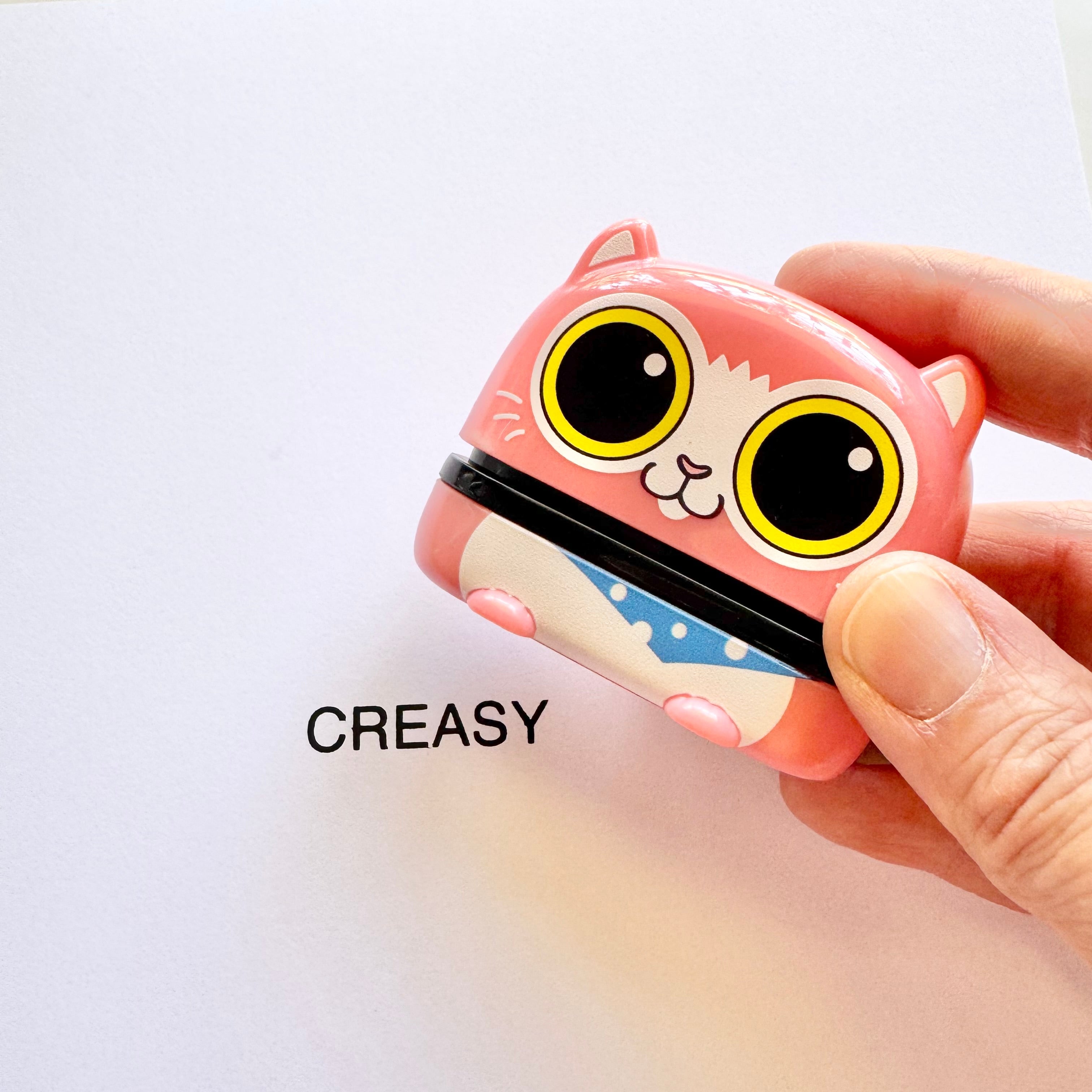 A person holding an animal name stamp with a cat printed on it above a sheet of paper with 'CREASY' printed on it