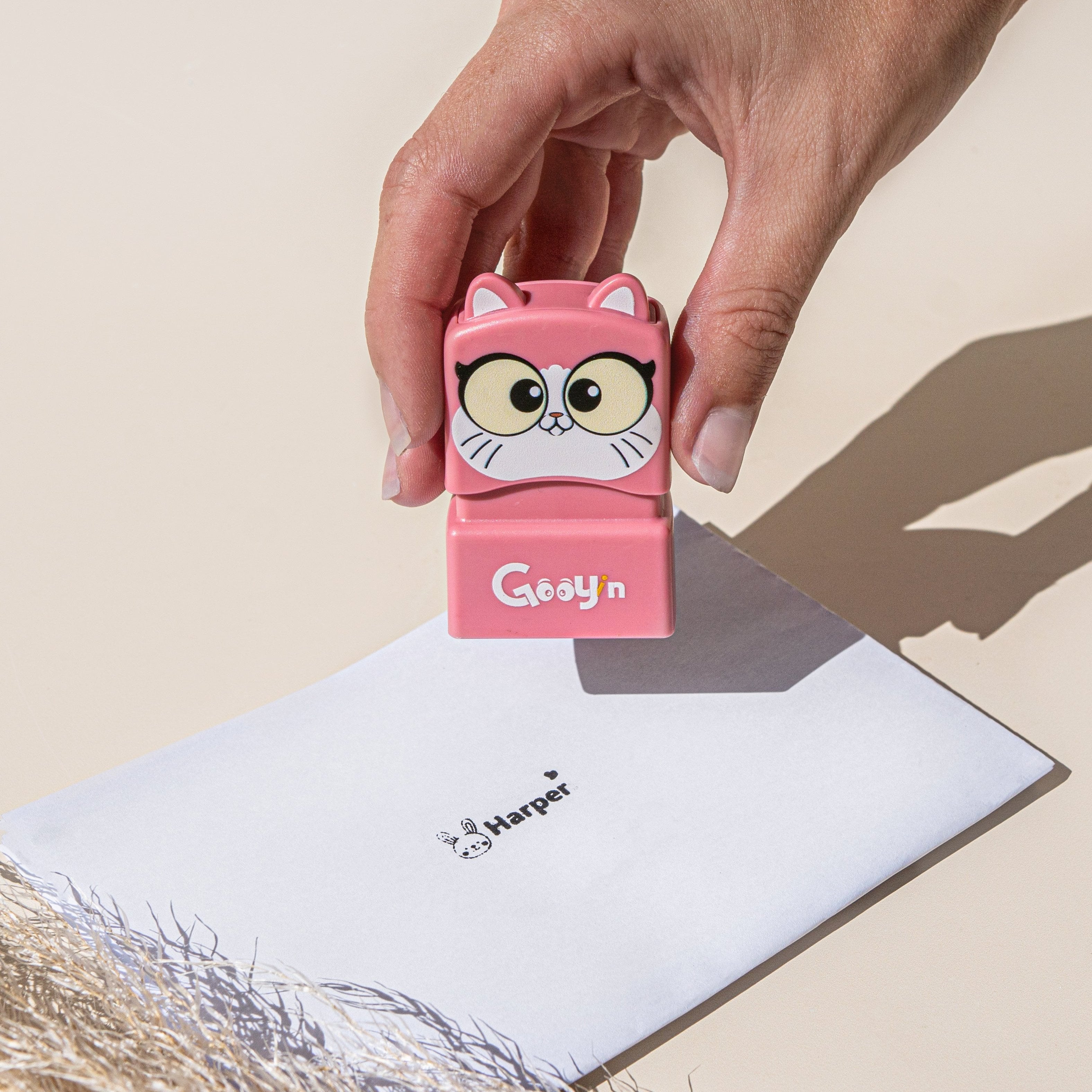 A person holding a cat name stamp with 'Gooyin' printed on the front above a sheet of paper with 'Harper' printed on it with a rabbit at the start and a heart at the end