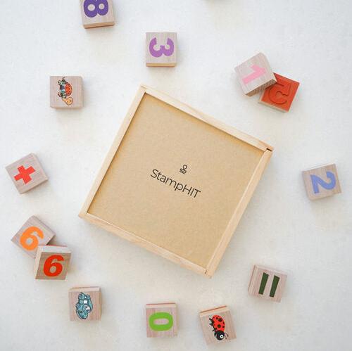 A wooden square with StampHIT printed on the middle surrounded by square education stamps with numbers, shapes and animals on them