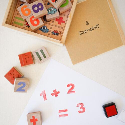 A top down view of a box of education stamps with numbers and shapes on them laying next to a sheet of paper with prints that say '1+2=3'. A sheet with StampHIT printed on it sits next to the box