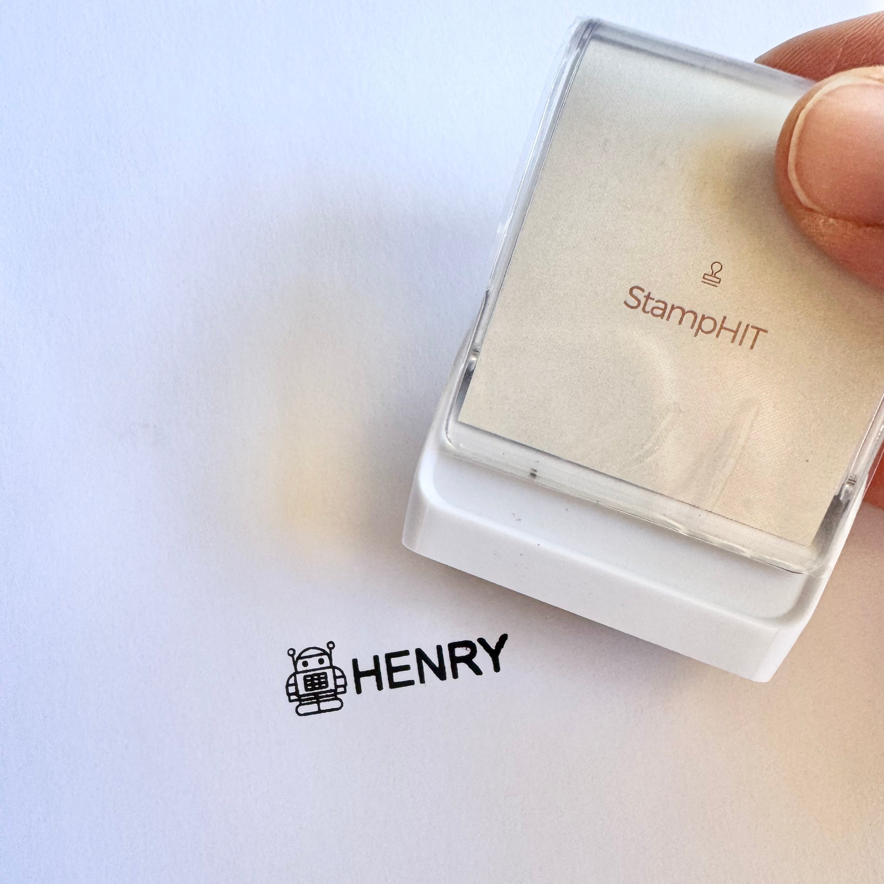 A white, rectangular StampHIT clothing name stamp being held over a sheet of paper with 'HENRY' printed over it with a robot at the start