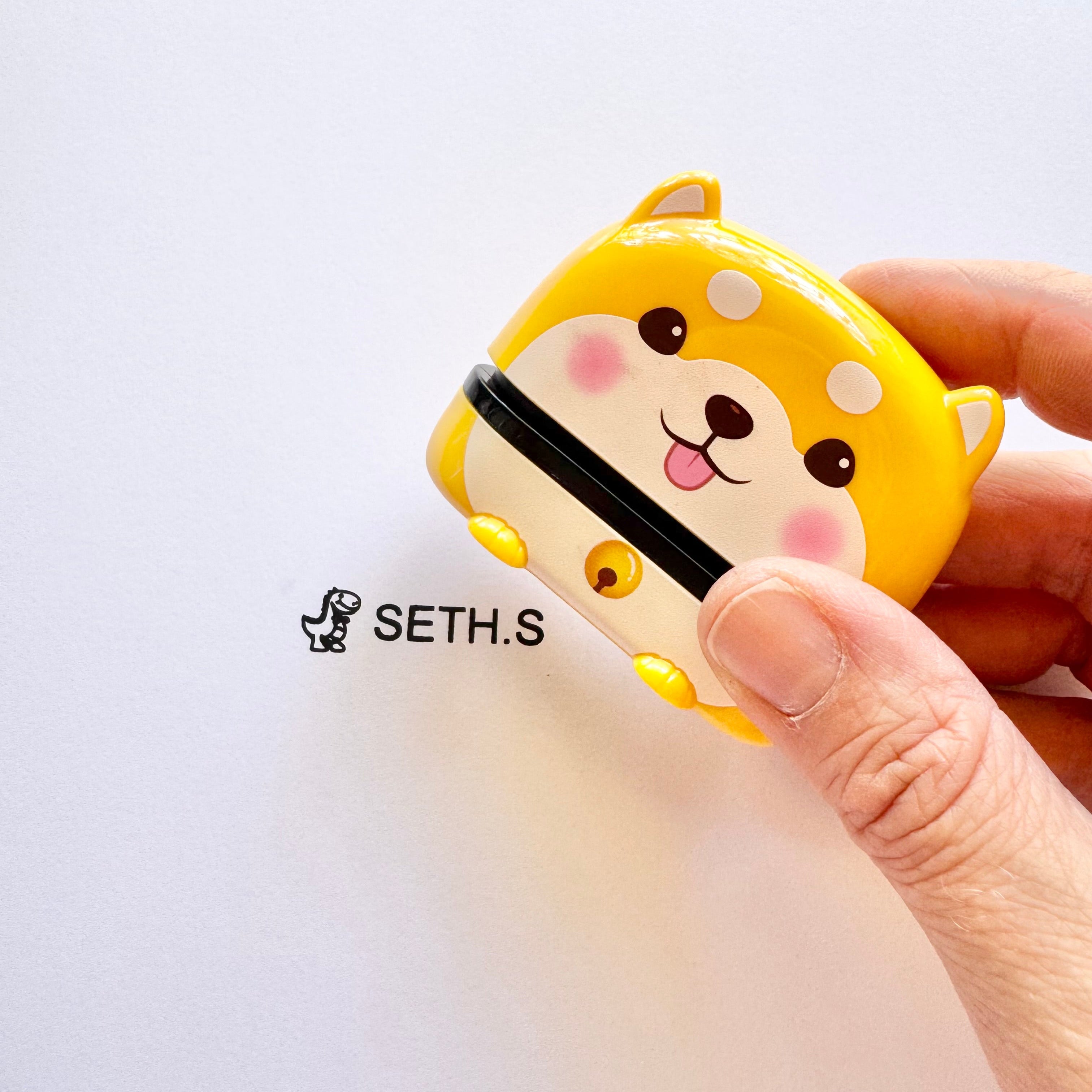 A person holding an animal name stamp that displays a dog over a sheet of paper with 'SETH.S' printed on it with a dinosaur at the start
