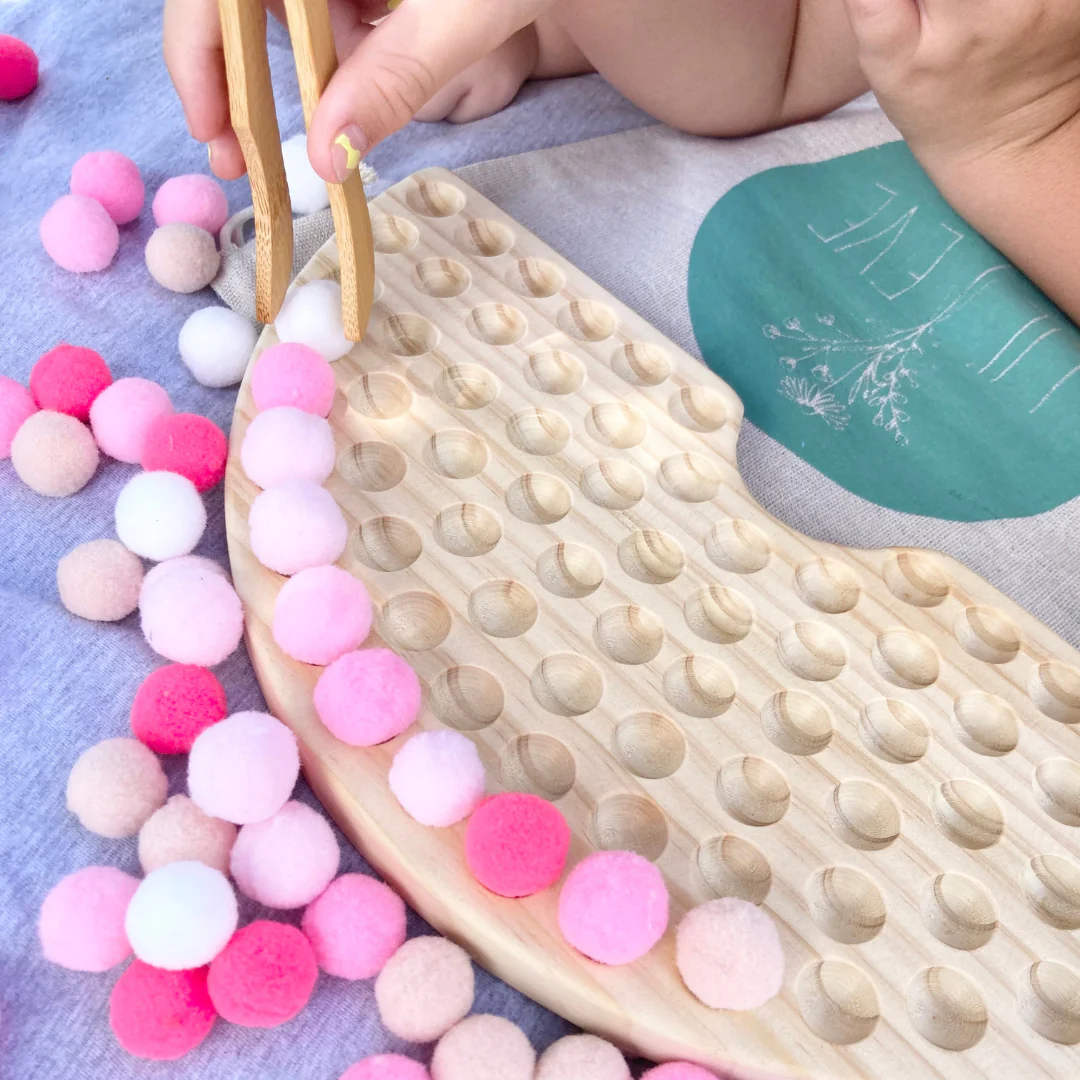 A child's hand, holding a wooden tong, placing coloured soft balls on a wooden board