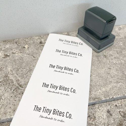 A grey business stamp with a rectangular base sitting next to a sheet of paper with 'The tiny bites co., Handmade to order' printed on it