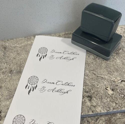 A grey business stamp with a rectangular base sitting next to a sheet of paper with A dreamcatcher and text displaying 'Dream catchers By Ashleigh' next to it