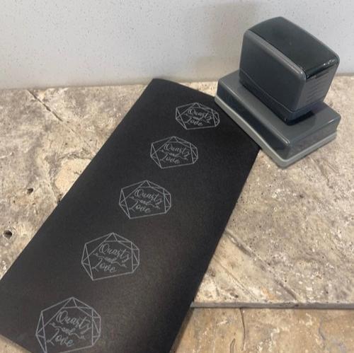 A grey business stamp with a rectangular base sitting next to a black piece of paper with diamonds and text displayed vertically along the length of the paper