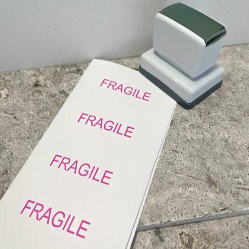 A white business stamp with a rectangular base sitting next to a sheet of paper with 'FRAGILE' displayed vertically along the length of the paper