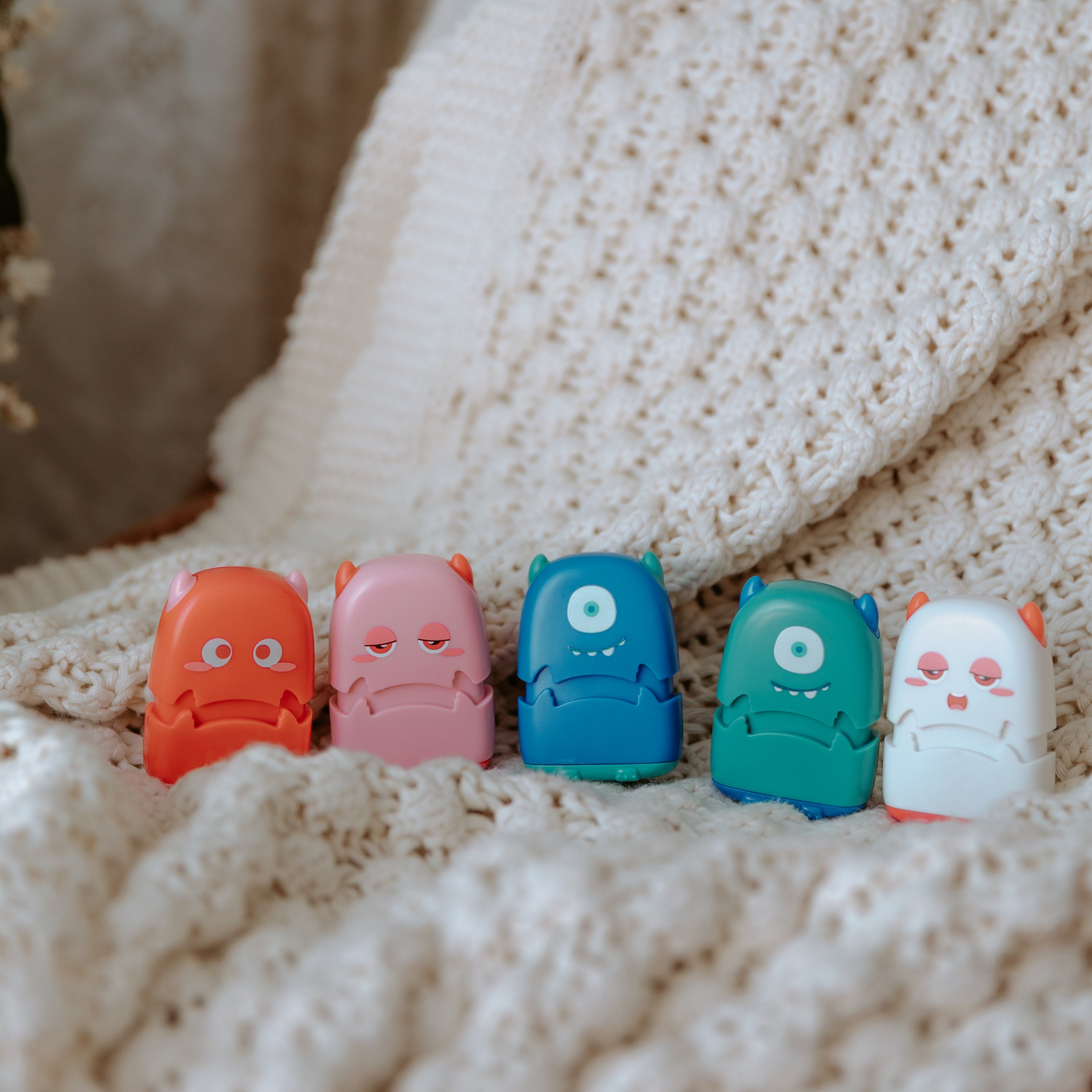 Five monster fabric name stamps sit upright on a woven cloth facing the camera. A red one, pink one, blue one, green one and a white one
