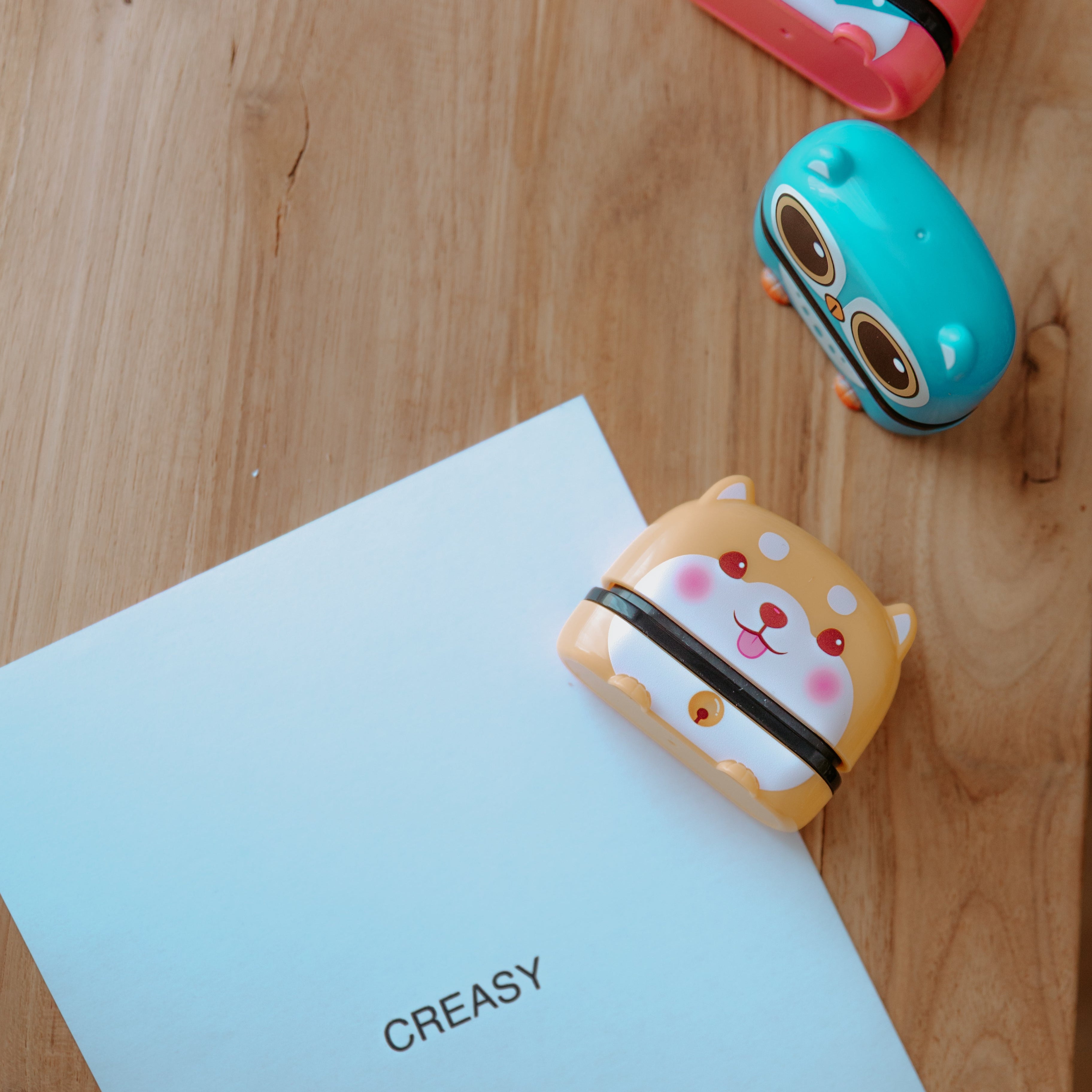 An animal name stamp laying on a sheet of paper with 'CREASY' printed on it. Another stamp displaying a bird is standing upright behind the dog and another stamp lays out of focus behind the bird