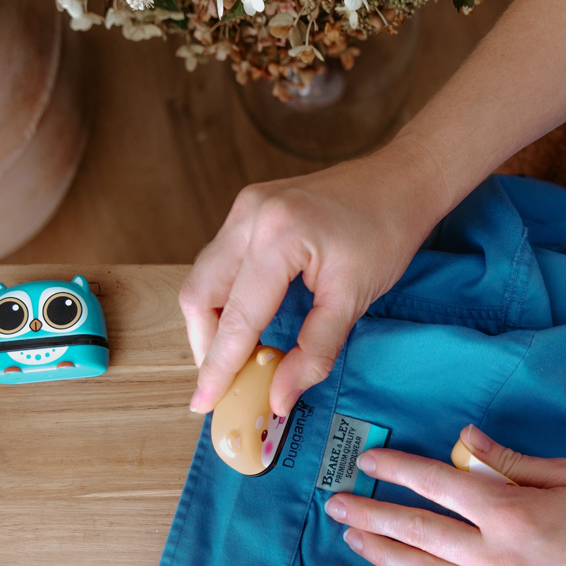 A person using an animal name stamp displaying a dog on a blue piece of clothes with another stamp displaying a bird laying on the table beside it. The print on the clothes displays 'Duggan' with a dinosaur at the end