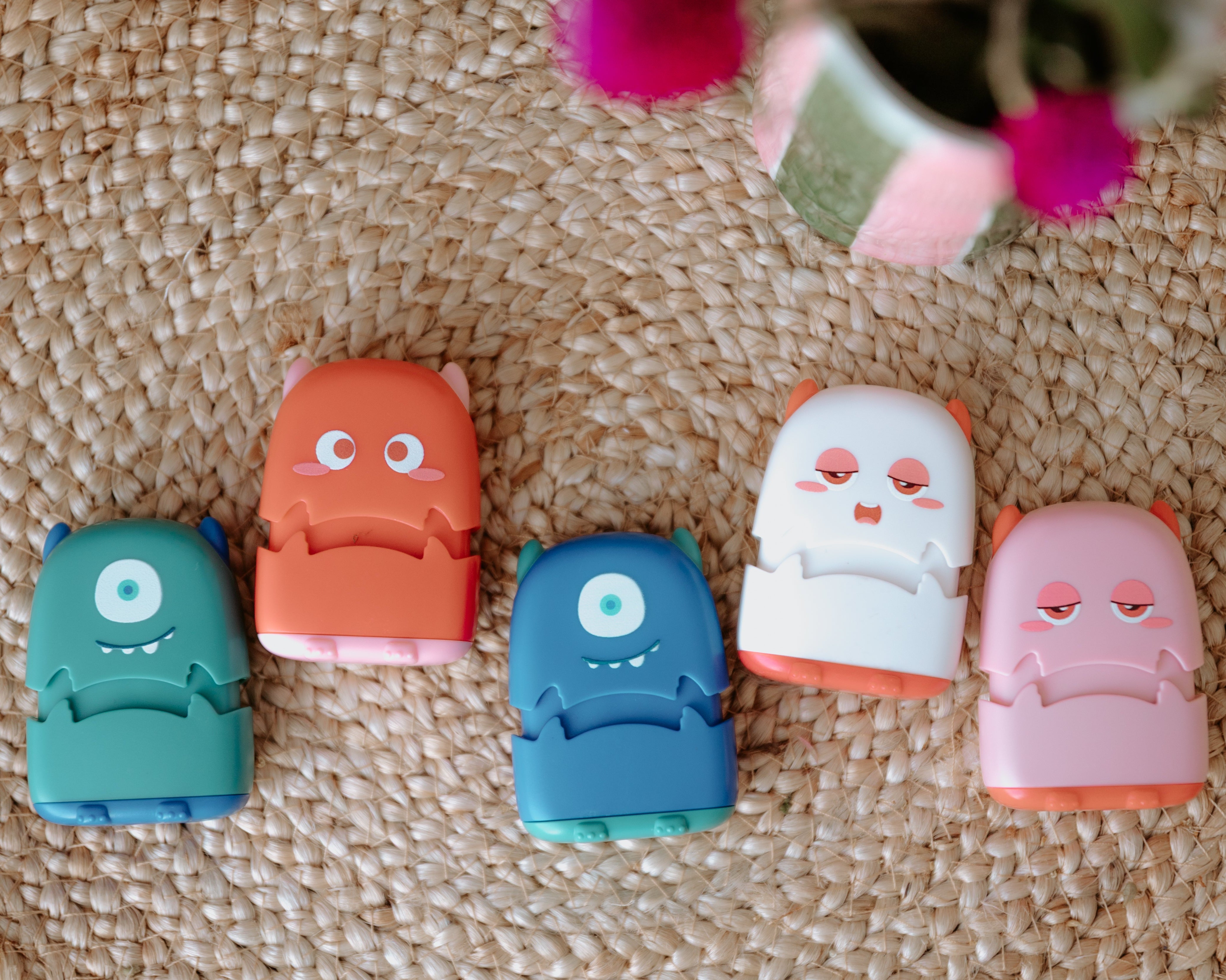 Five monster fabric name stamps lay face up on a woven rug. A green one, red one, blue one, white one and pink one