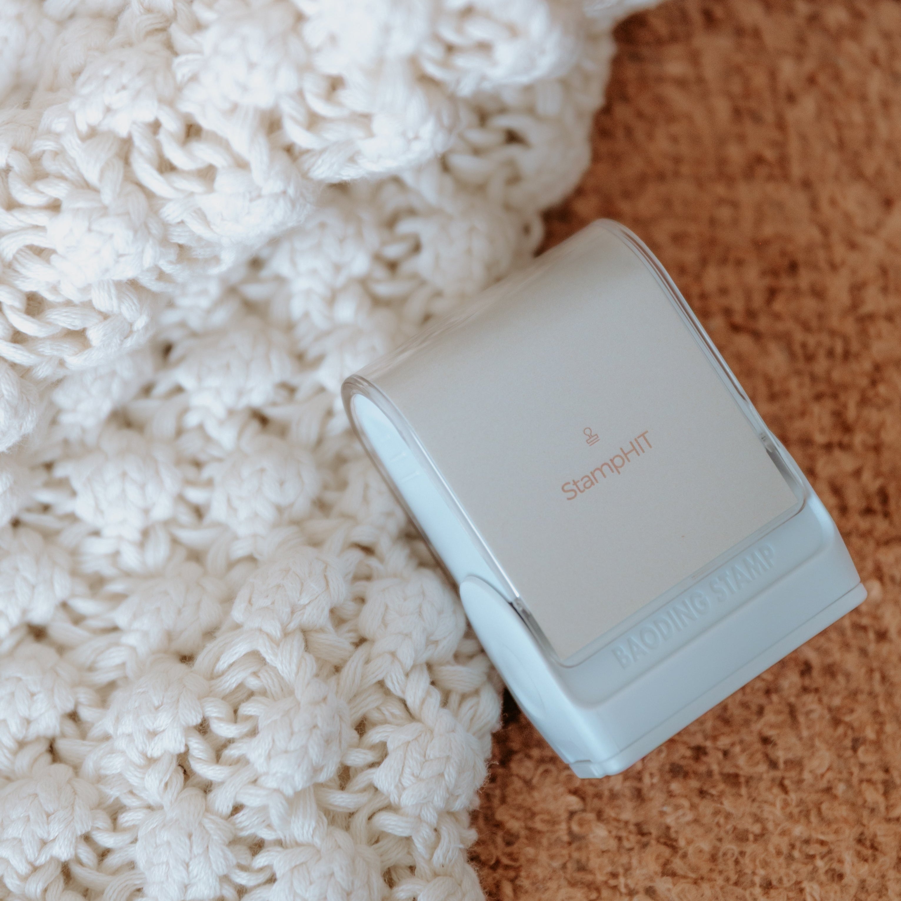 A white rectangular clothing name stamp with a curved top, leaning on a woven cloth displaying the StampHIT logo