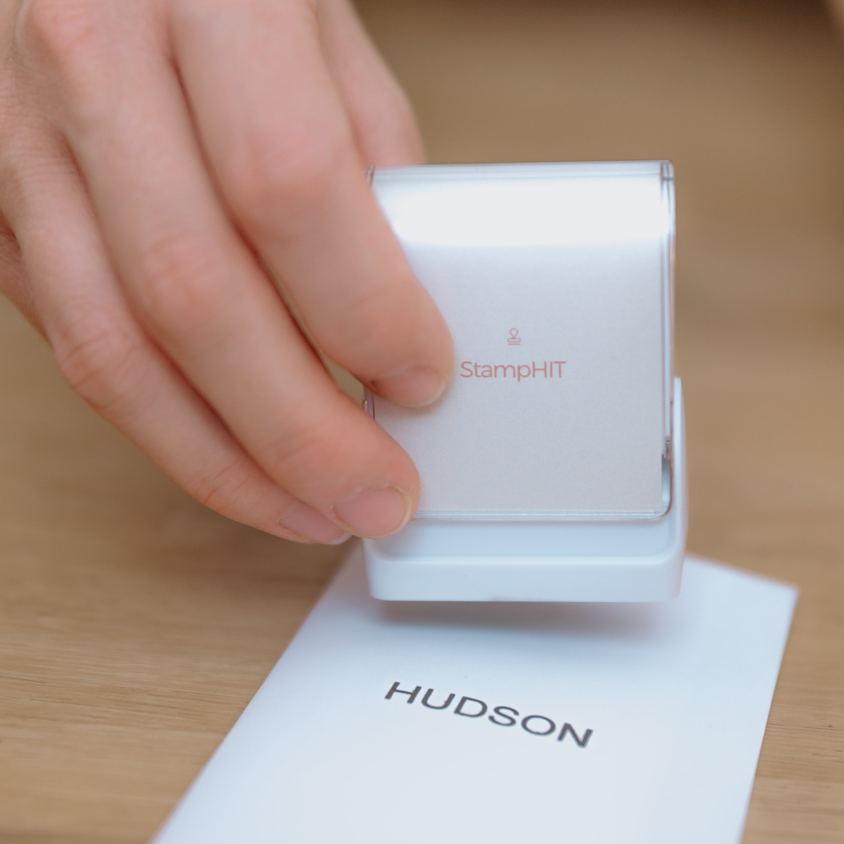 A person holding a white, rectangular clothing name stamp with a curved top displaying the StampHIT logo on the front of the stamp over a piece of paper with 'HUDSON' stamped on it