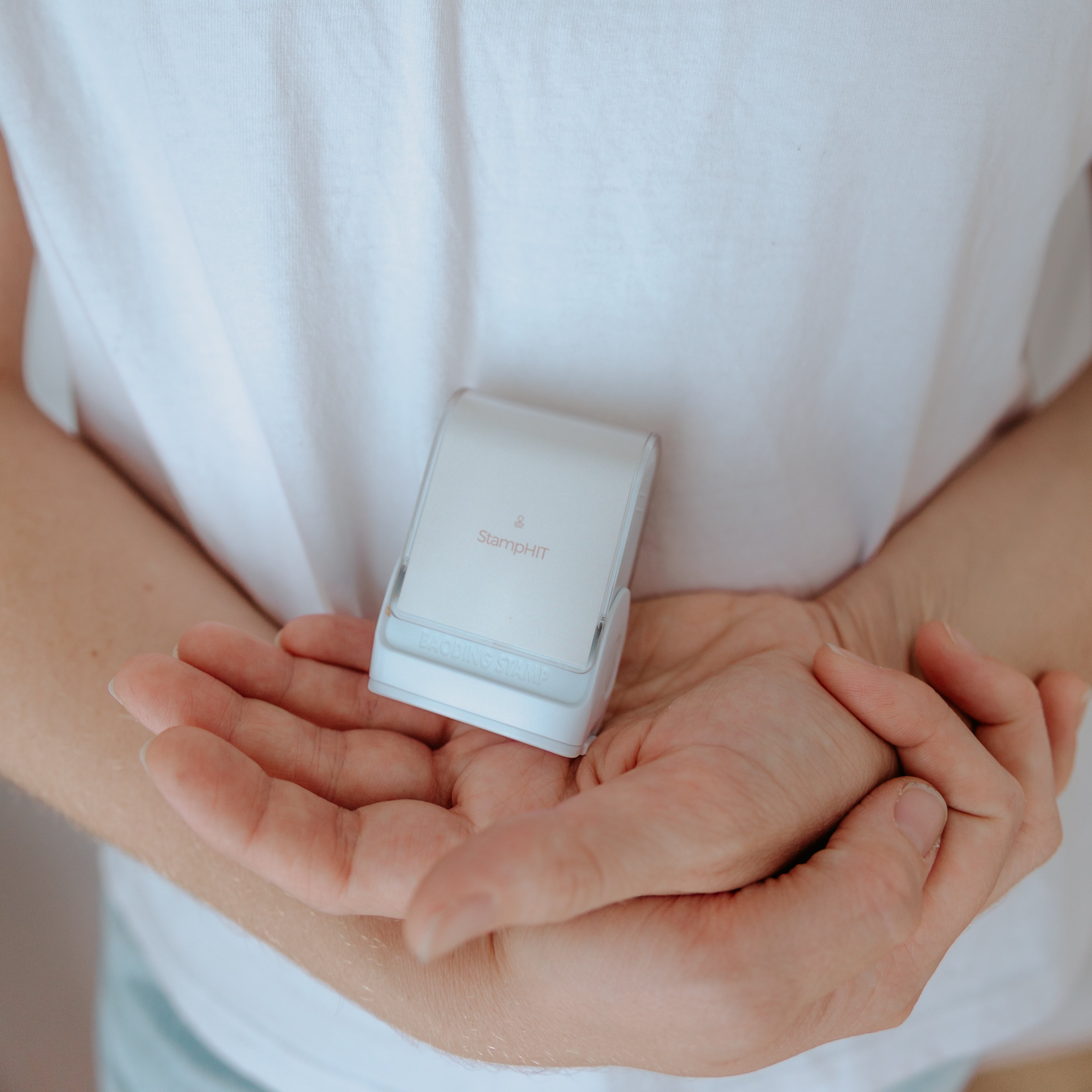 A person wearing a white shirt holding a white rectangular clothing name stamp with a curved top displaying the StampHIT logo printed on the front of the stamp in the palm of their hand