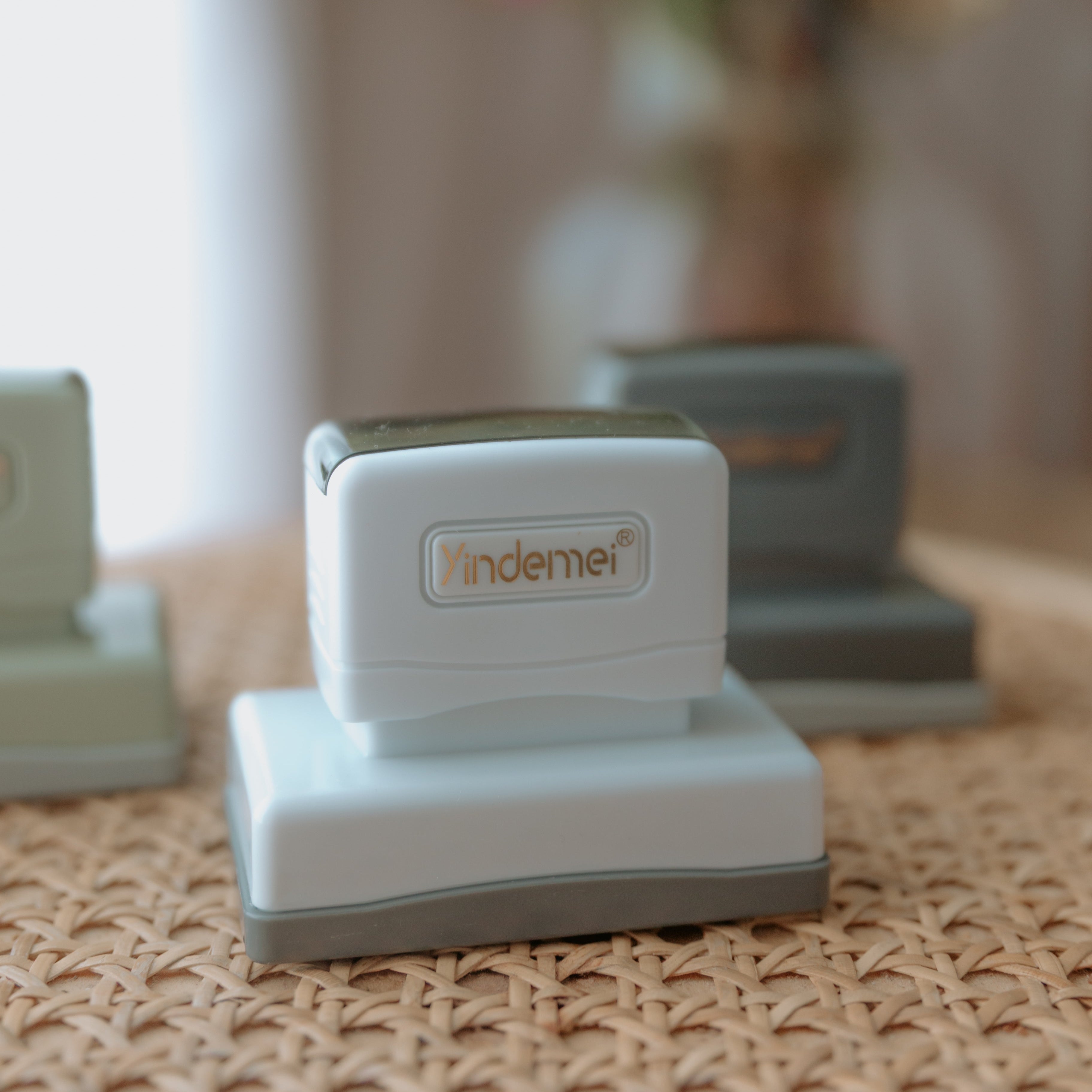 A white business stamp with a squared base and text that displays 'Yindemei' facing the camera with two stamps out of focus behind it
