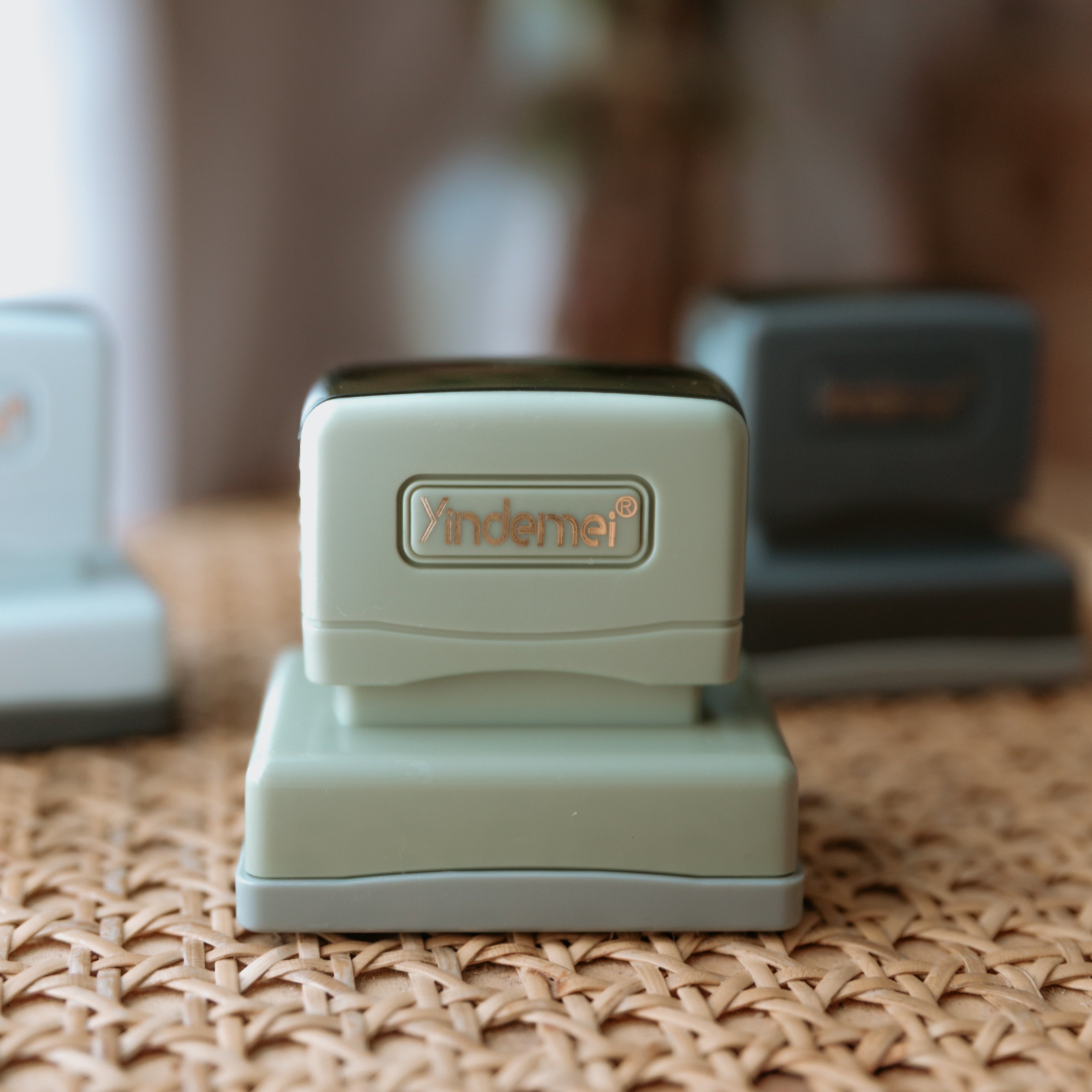 A pale grey business stamp with a squared base with text that displays 'Yindemei' facing the camera with two stamps out of focus behind it