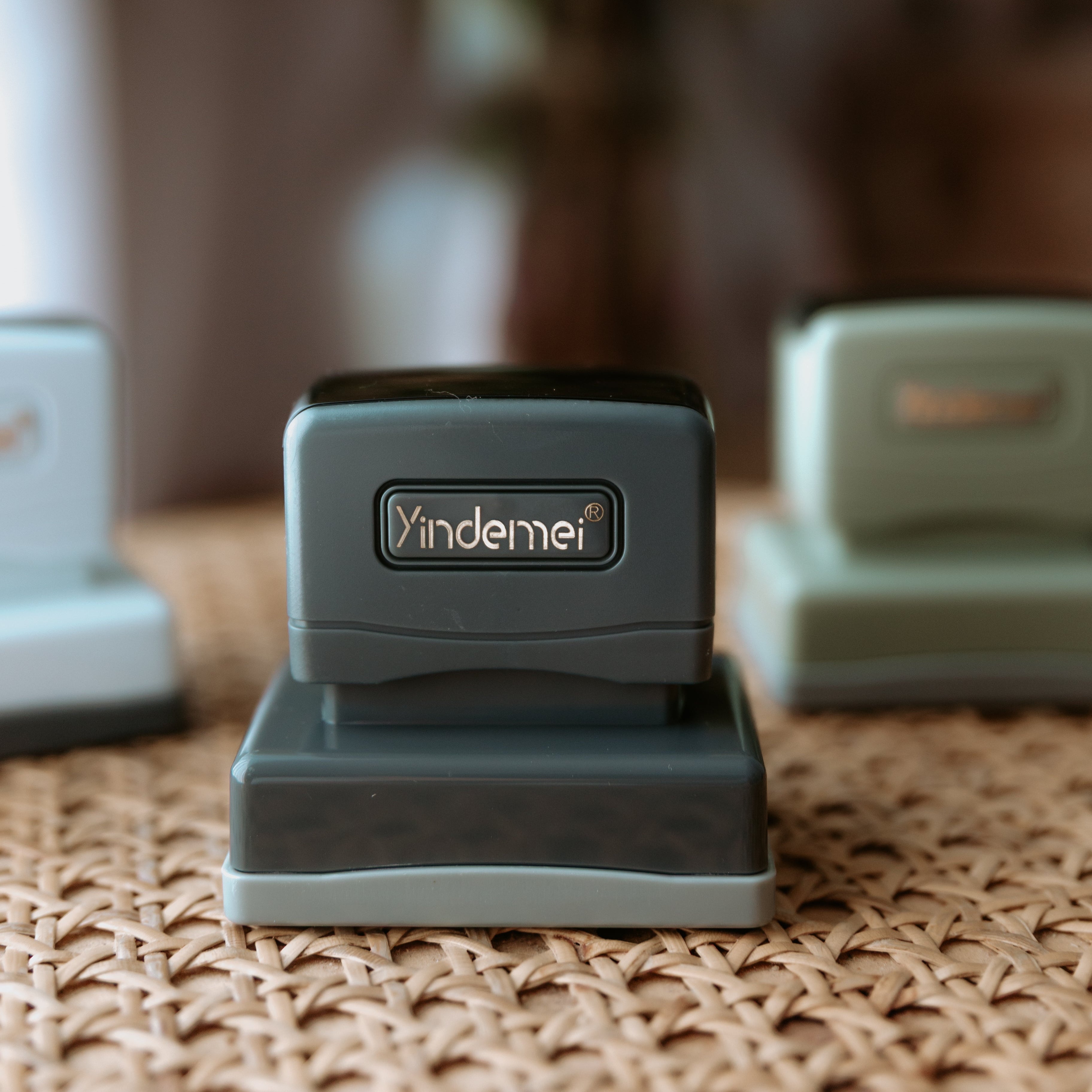 A dark grey business stamp with a squared base facing the camera with text that displays 'Yindemei' and two other stamps out of focus behind