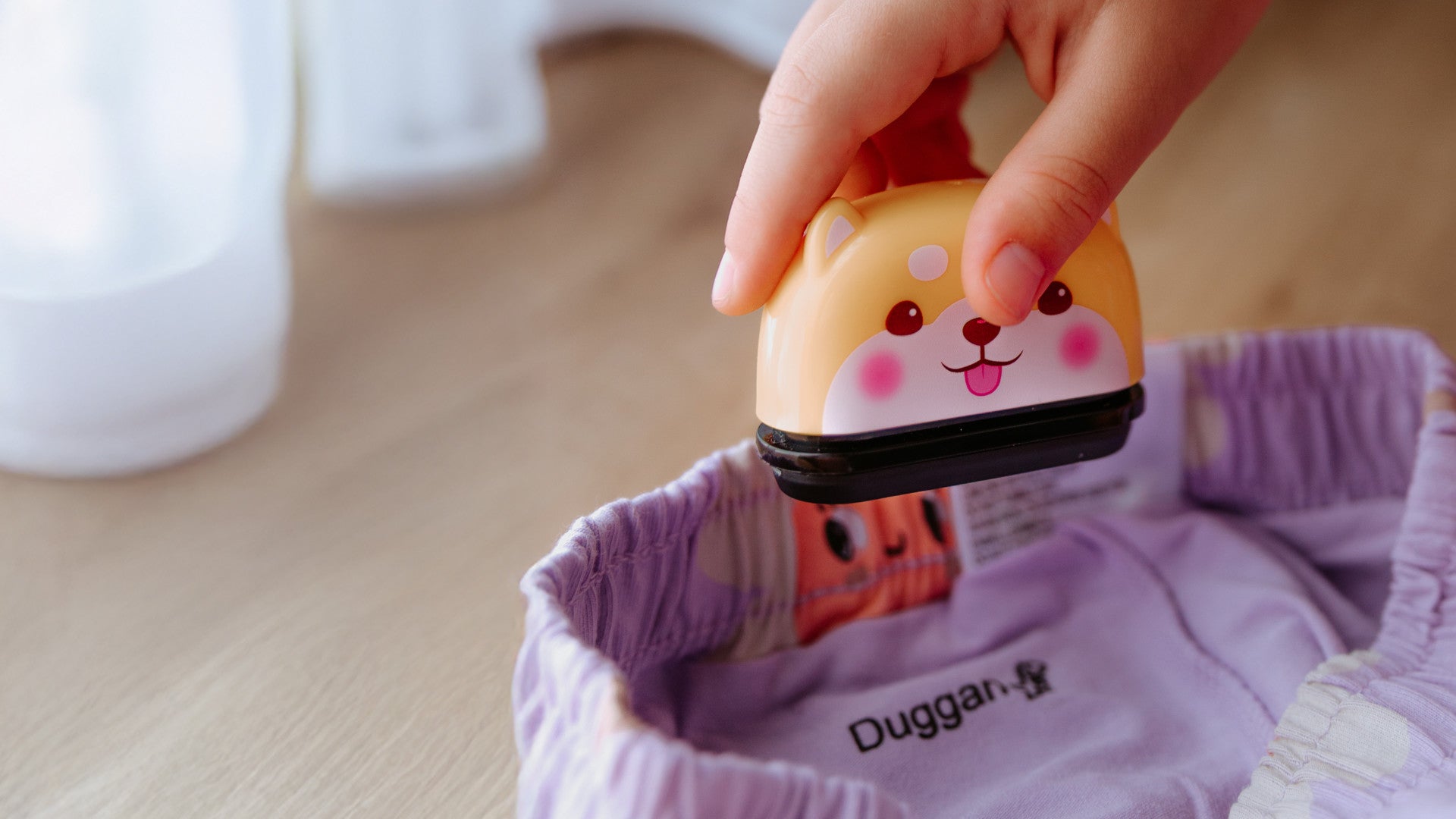A hand holding a StampHIT Fabric Ink stamp with a cute Bear face stamps the name Duggan on a purple shirt collar.