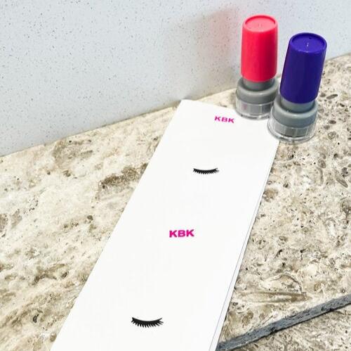 A red and blue card stamp next to a sheet of paper with 'KBK' and eyelashes in a vertical pattern